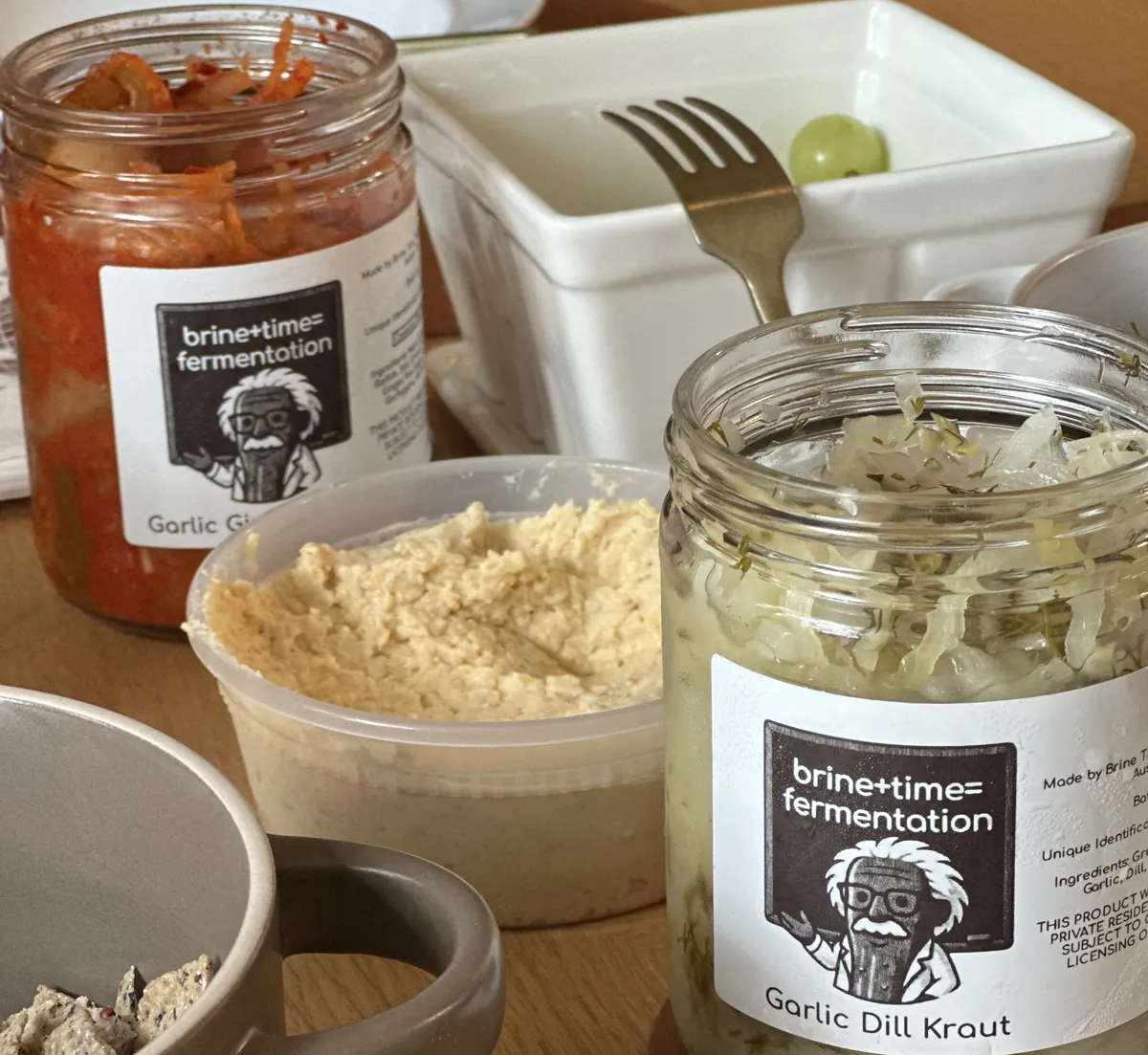 Open jars of Brine Time garlic ginger kimchi and garlic dill sauerkraut on a table next to a tub of fermented hummus and tortilla chips